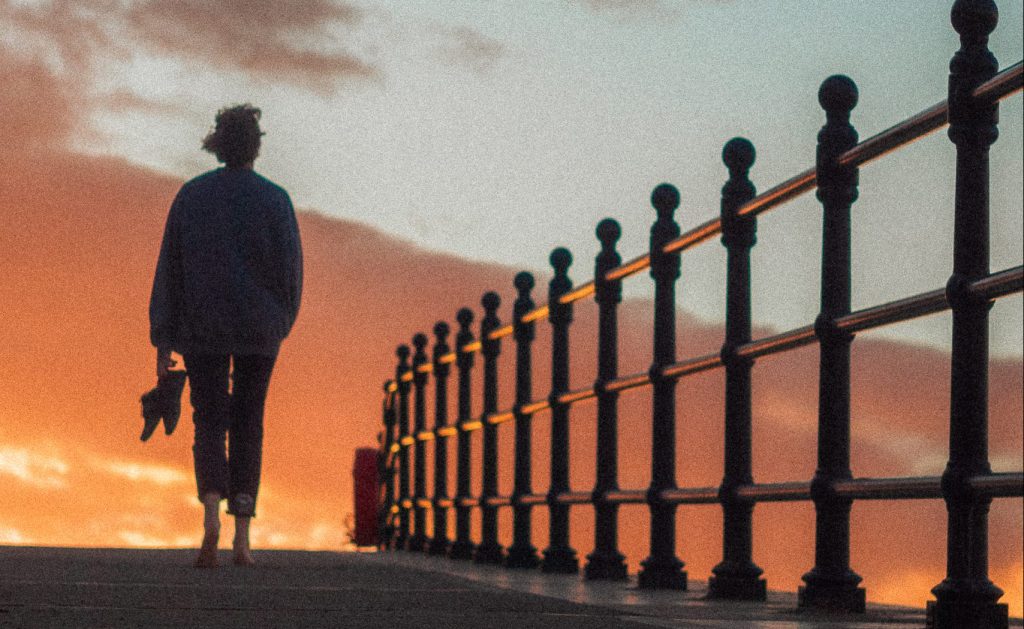 A silhouette of a figure walking towards a golden sunset, beside them is a silhouette of railings.