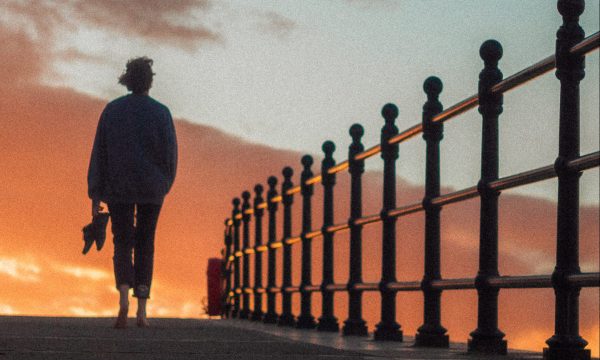 A silhouette of a figure walking towards a golden sunset, beside them is a silhouette of railings.