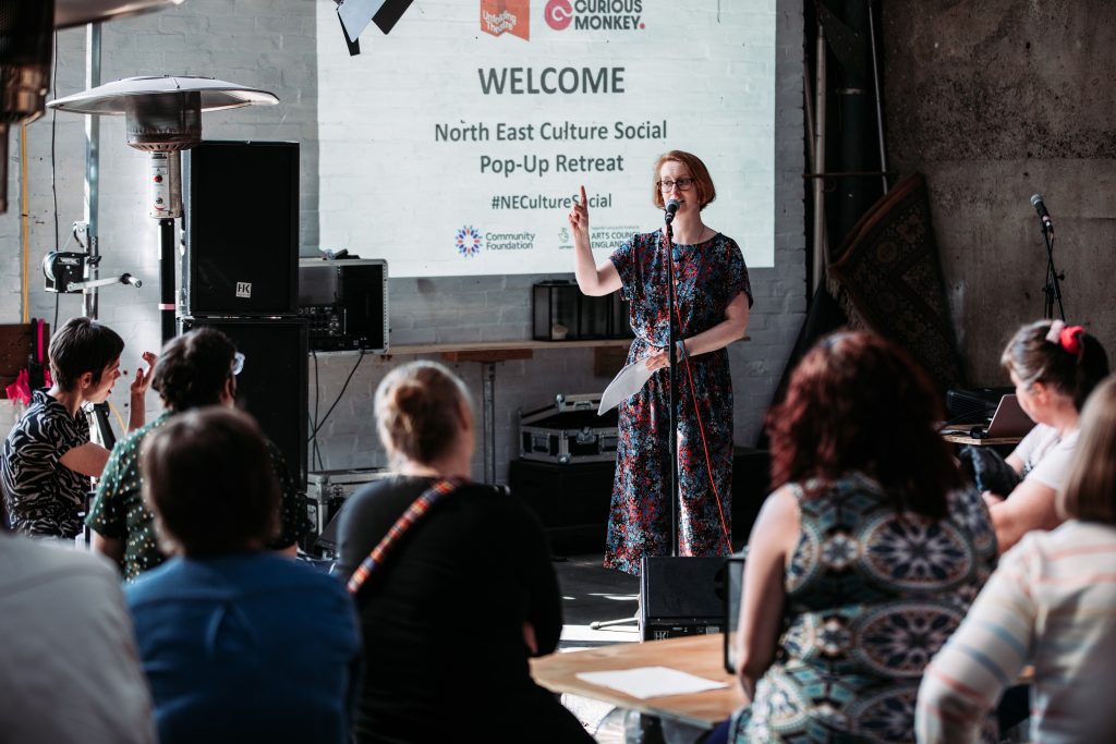 A white woman with short hair and glasses is standing behind a microphone, speaking to a seated audience in front of her. A screen behind her shows the details of this previous event and reads 'WELCOME North East Culture Social Pop-Up Retreat #NECultureSocial