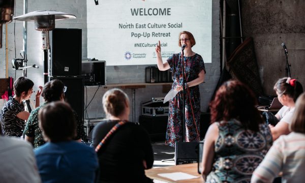 A white woman with short hair and glasses is standing behind a microphone, speaking to a seated audience in front of her. A screen behind her shows the details of this previous event and reads 'WELCOME North East Culture Social Pop-Up Retreat #NECultureSocial