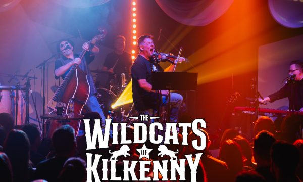 Mike from Wildcats plays fiddle on stage to a busy crowd, there's a double bass player, keyboard player and drummer also on stage with Mike