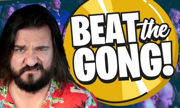 Matt Reed, a white comedian with mid-length dark brown hair, stands next to an animated Beat the Gong logo. He is looking directly into the camera and is wrinkling up his nose.