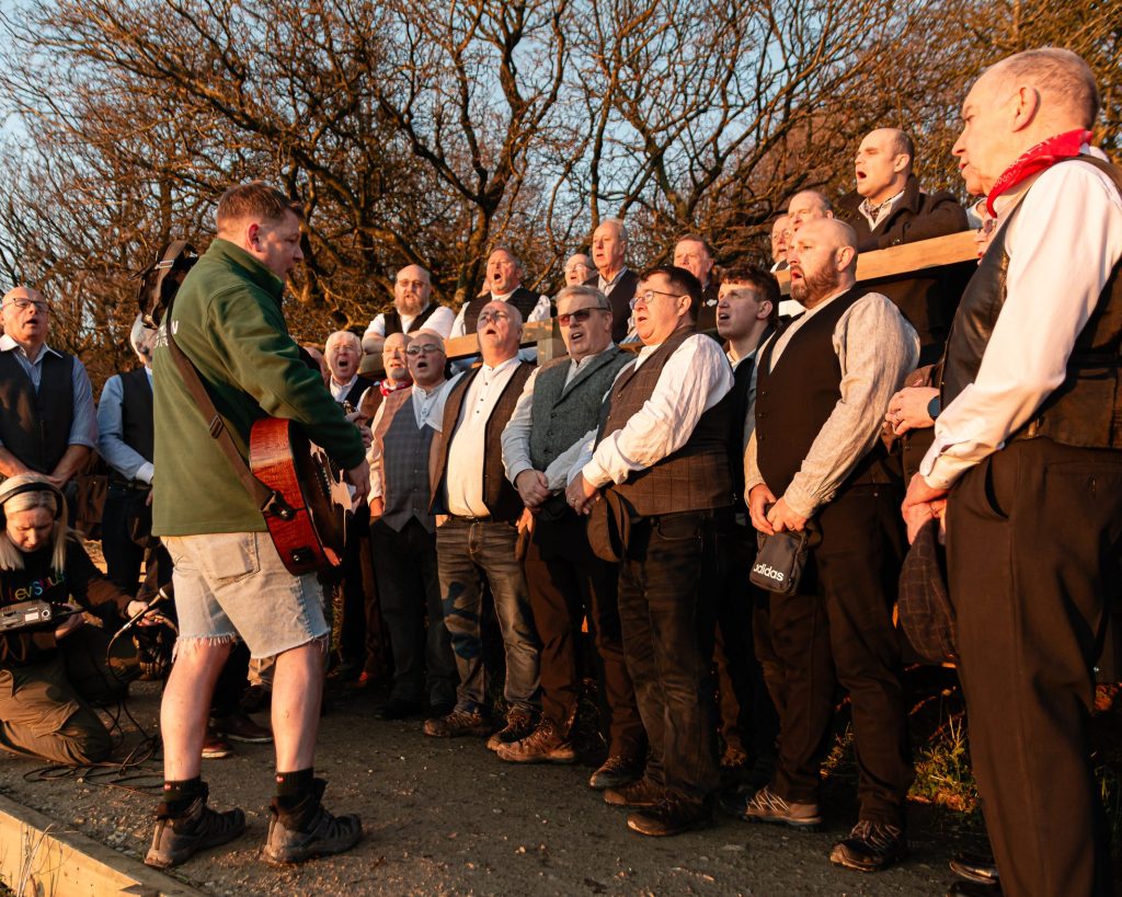 Infant Hercules male choir stand singing around Mike McGrother with his acoustic guitar