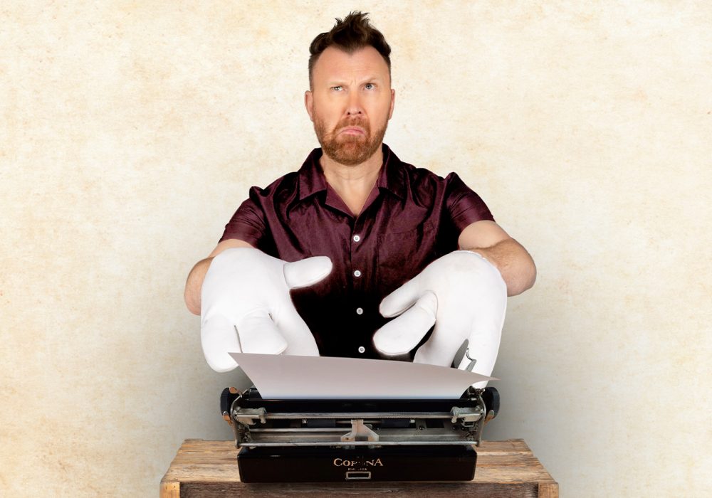 Comedian Jason Byrne, a white middle-aged man with short brown hair, is sitting in front of a typewriter, he is wearing comically large white gloves and a confused, thoughtful expression.