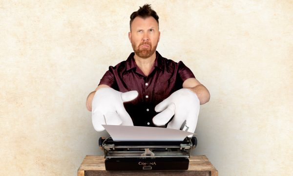 Comedian Jason Byrne, a white middle-aged man with short brown hair, is sitting in front of a typewriter, he is wearing comically large white gloves and a confused, thoughtful expression.