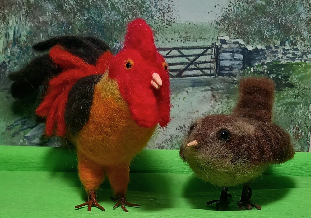 Two needle felted birds stand in front of a farmyard scene. On the left is a large cockerel, and on the right is a small, round, sparrow.