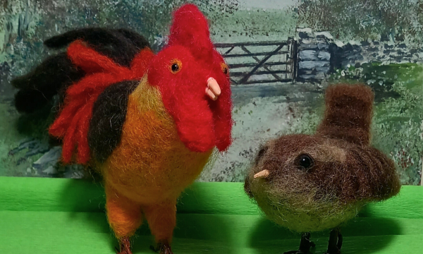 Two needle felted birds stand in front of a farmyard scene. On the left is a large cockerel, and on the right is a small, round, sparrow.
