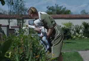 A woman holding a baby outside in a garden who is reaching out to touch a plant.