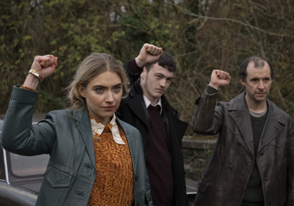 Image of Imogen Poots raising one fist in the air while looking past the camera, two men stand in the background behind her also raising one fist in the air.