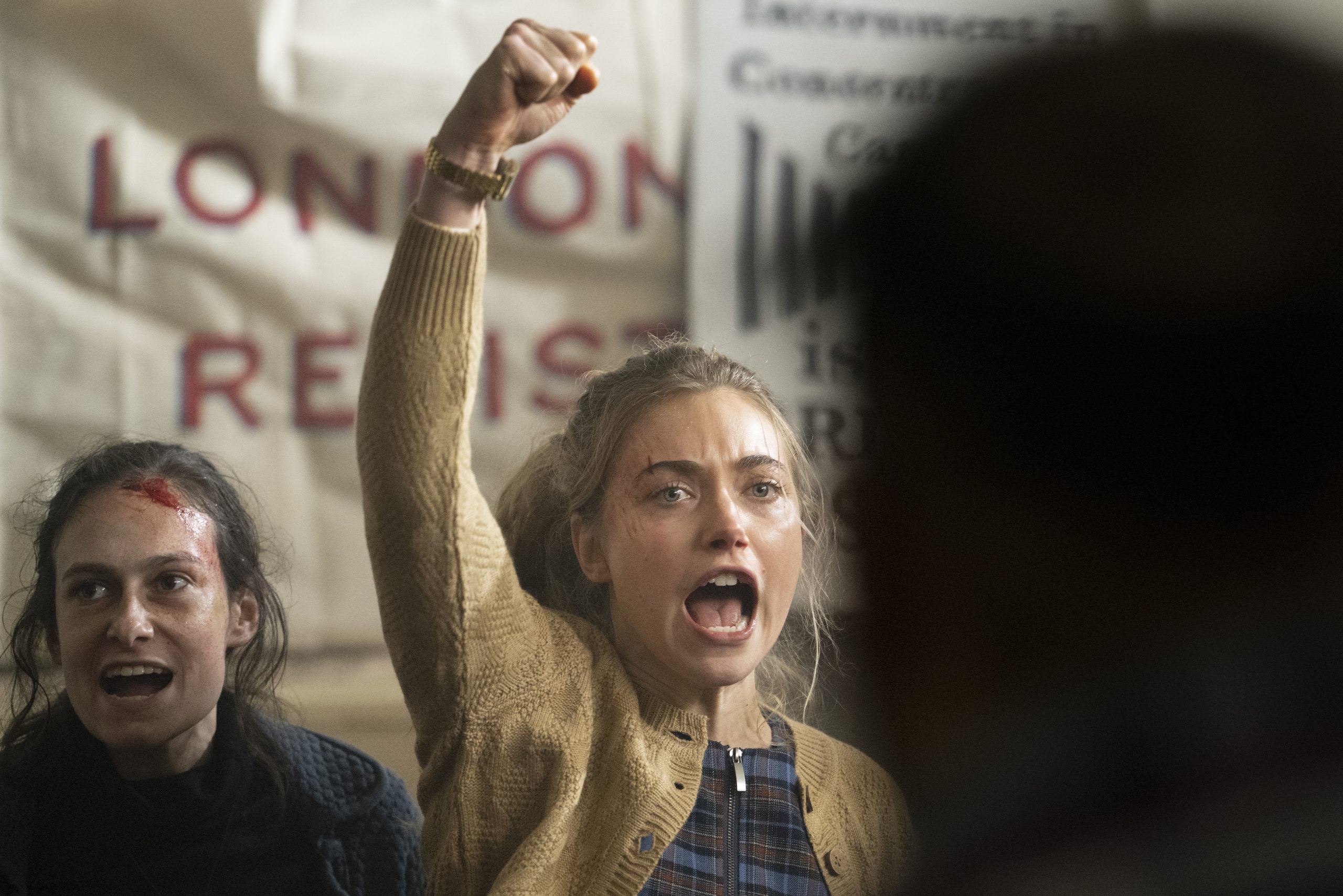 Imogen Poots stands with one hand raised high above her head, in the background there is woman with a bloody head and banners raised.