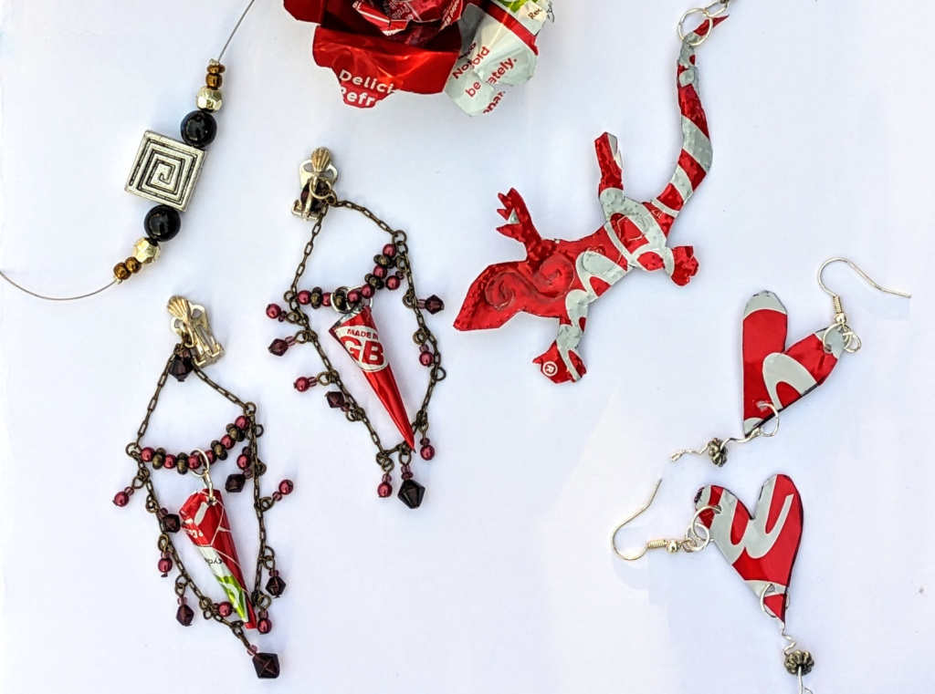 Jewellery created by recycled coca cola cans. There is a pendant shaped like a lizard, a pair of dangly, beaded earrings, and a pair of earrings with heart-shaped pendants hanging from them.