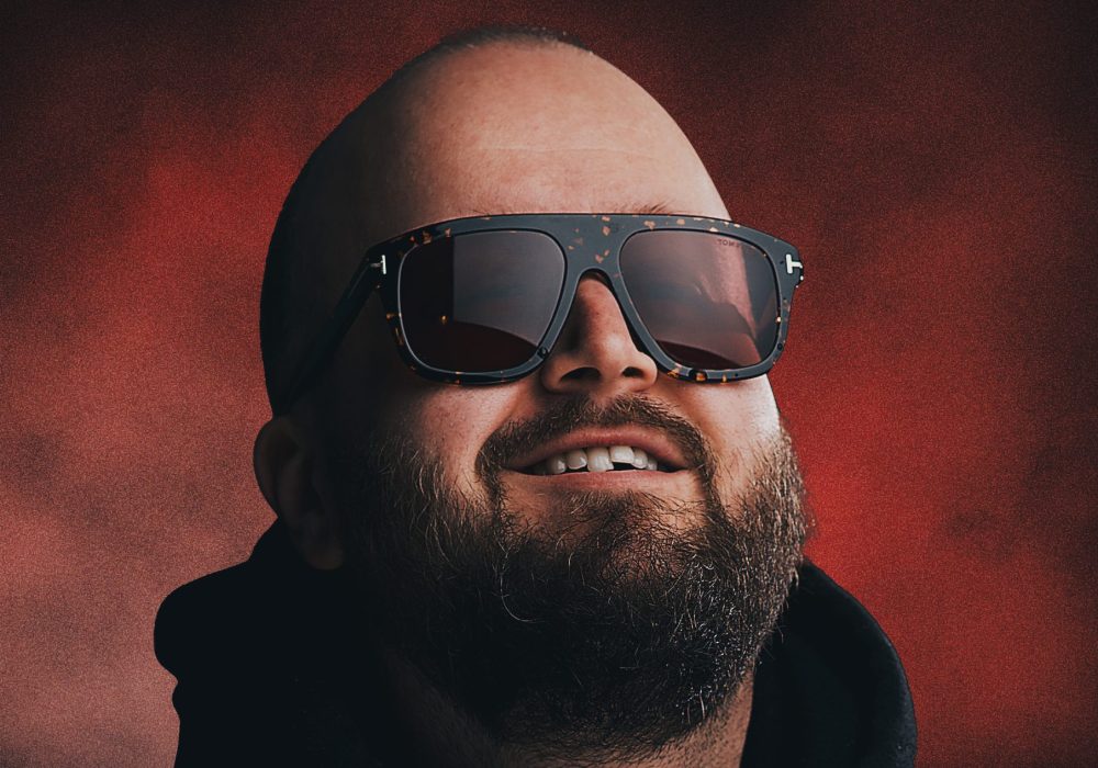 A headshot of comedian Freddy Quinne, a white man with a bald head and a beard, he is grinning and wearing dark sunglasses.