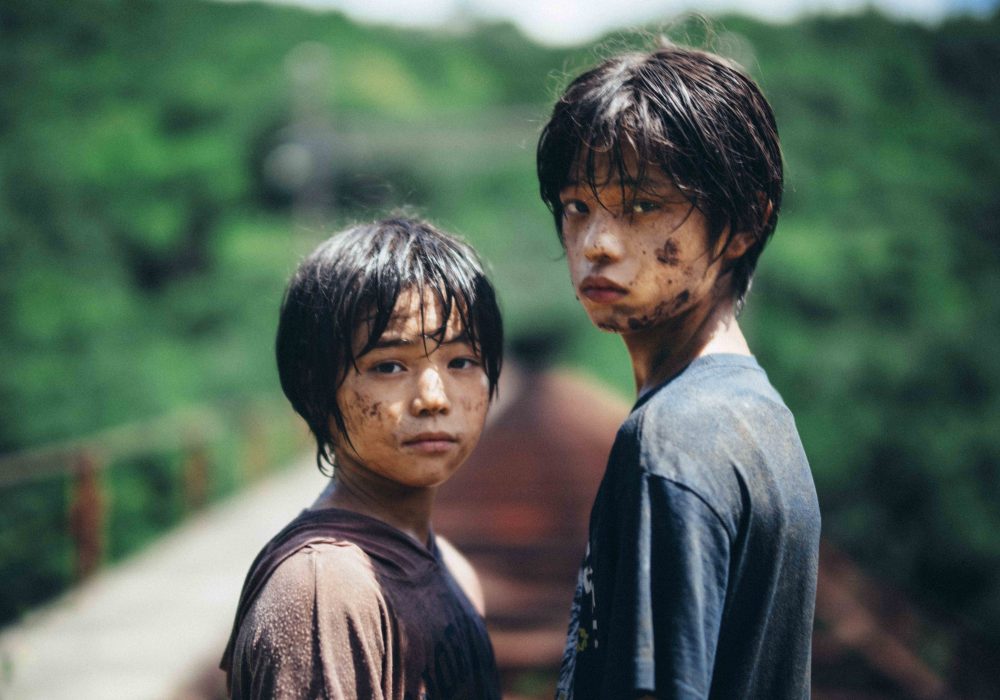 Two Japanese boys stand covered in mud facing directly into the camera lens. They are covered in mud, in the blurred background we can see railway tracks running away from them and trees on the horizon.