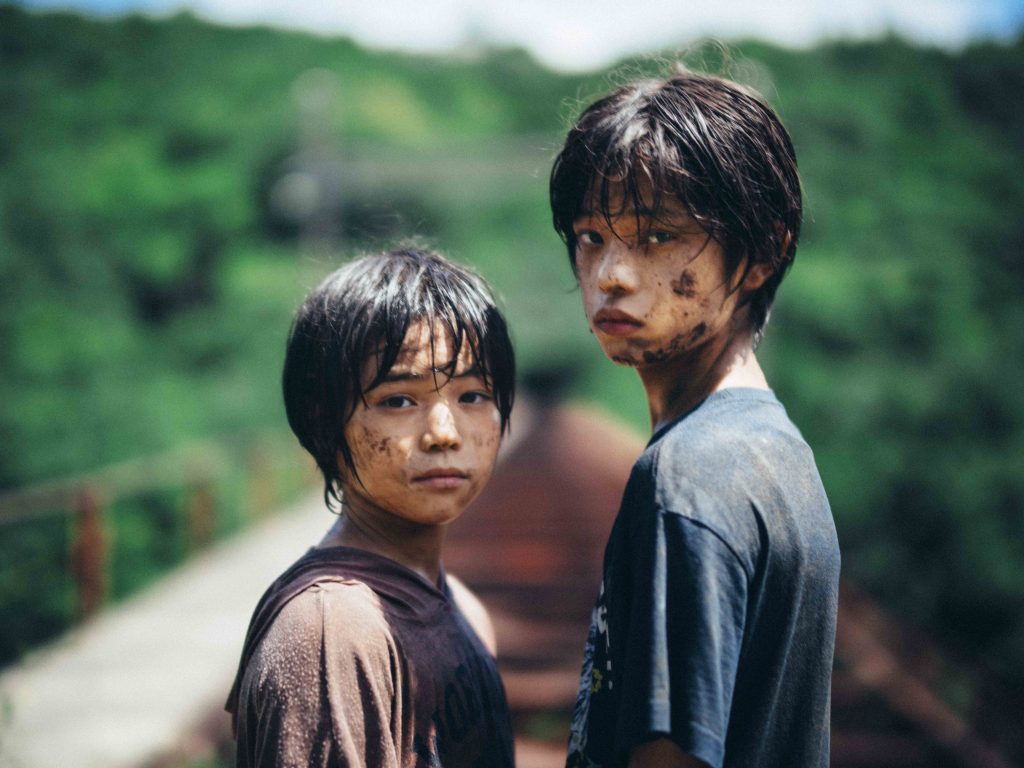 Two Japanese boys stand covered in mud facing directly into the camera lens. They are covered in mud, in the blurred background we can see railway tracks running away from them and trees on the horizon.