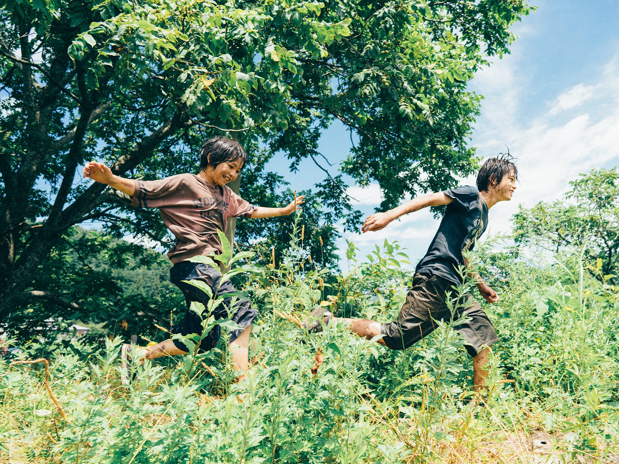 Two Japanese boys covered in mud run through the long grass smiling. Behind them is a large tree and a blue sky with wispy white clouds.