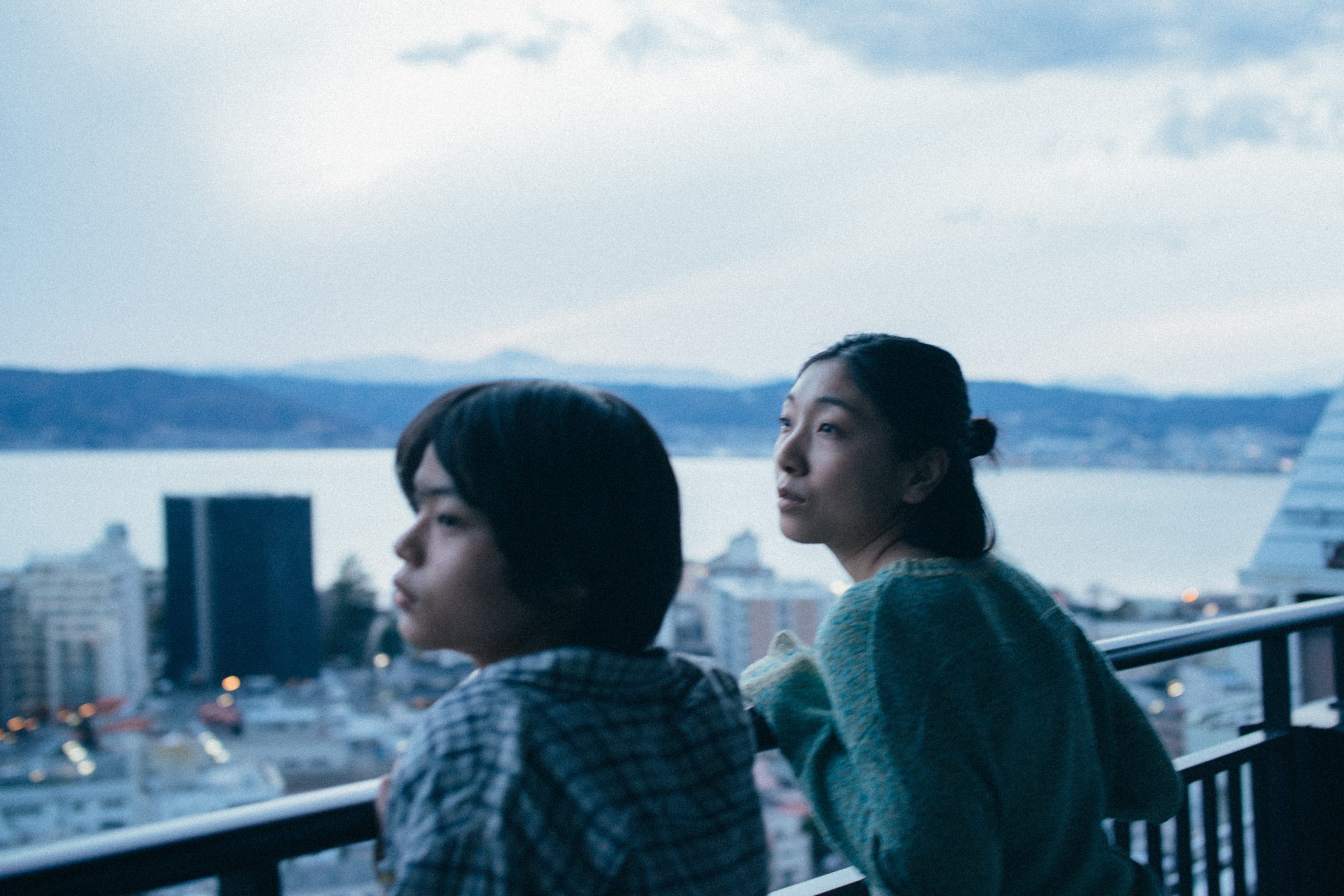 A Japanese boy and his mother stand looking out from their balcony with forlorn expressions. In the distance we can see the city, a body of water, and the mountains.