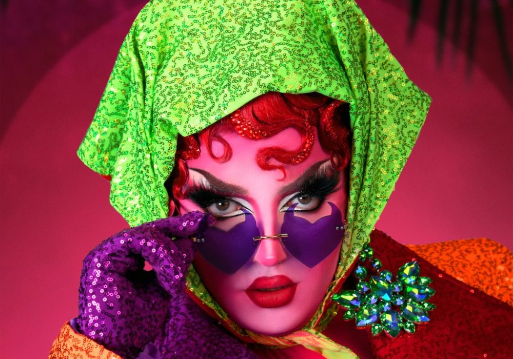 A headshot of drag queen Choriza May, she has pale pink skin, and is wearing a bright green headscarf, she has on a glittery purple glove and is looking over the top of purple heart shaped sunglasses.