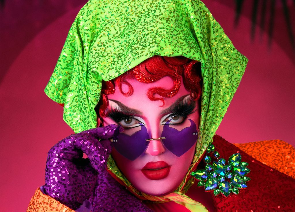 A headshot of drag queen Choriza May, she has pale pink skin, and is wearing a bright green headscarf, she has on a glittery purple glove and is looking over the top of purple heart shaped sunglasses.