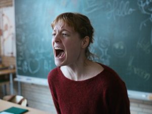 A white female who is a teacher stood in front of a chalk board. She is wearing a dark red top and her expression is screaming.