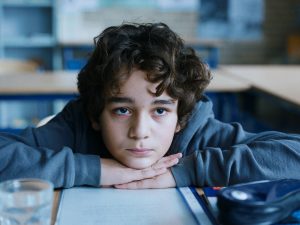 Image of a white Turkish child with short brown curly hair. Wearing a dark blue hoodie sat at a desk with his arms crossed resting his head on his hands.