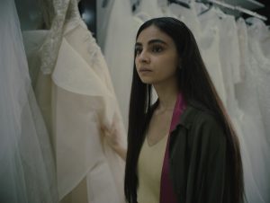 Female with long dark hair wearing a lemon top and khaki coat in a shop surrounded by wedding dresses.