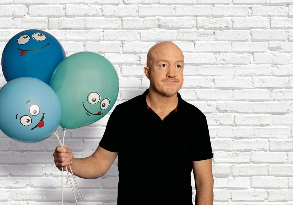 Comedian Andy Parsons, a white, middle-aged, bald man wearing a black shirt and standing in front of a white brick wall. He is holding 3 balloons in varying shades of blue, each with a cartoon face on.