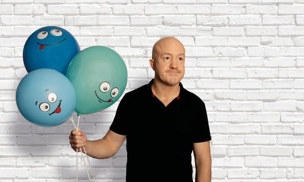 Comedian Andy Parsons, a white, middle-aged, bald man wearing a black shirt and standing in front of a white brick wall. He is holding 3 balloons in varying shades of blue, each with a cartoon face on.
