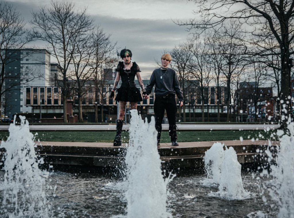 A fountain with 2 people stood behind the fountain holding hands. Both are dressed it black gothic clothing.