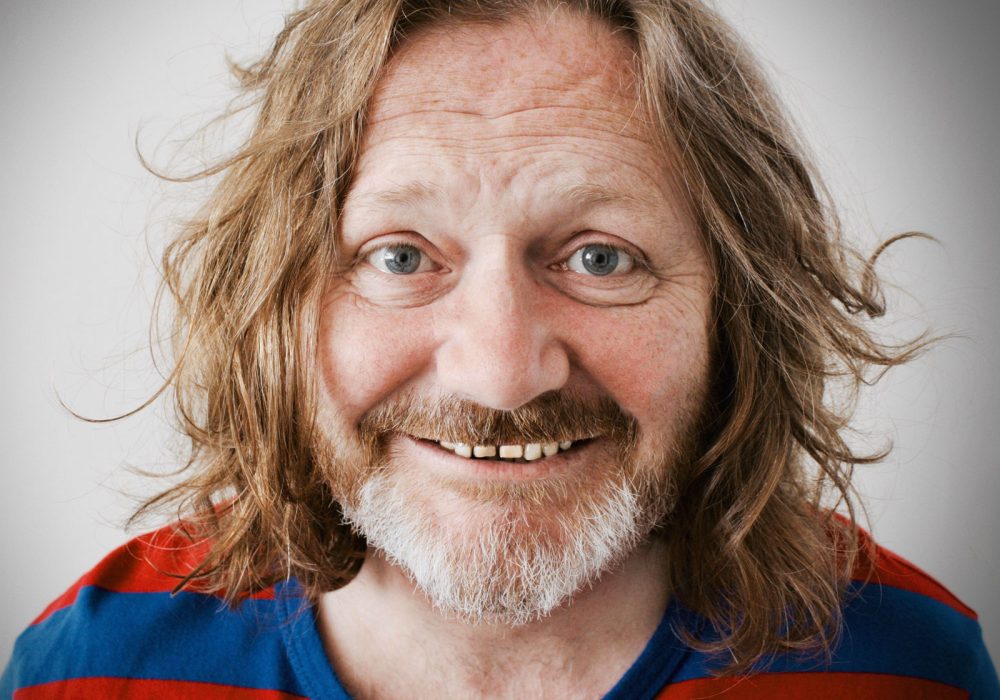 John Fothergill stands facing you, he has long wavy hair and a red and blue top on with a beard