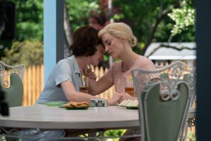 Two females sat at a garden table. One with blone hair and a pale pink dress and one with dark brown hair with a pale blue dress. Their heads together looking as if they are consoling each other.