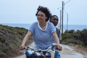 Two women riding a moped. The one driving has shoulder length dark curly hair wearing a blue and white striped tshirt. The other is wearing a pale blue tshirt and khaki backpack and has very short dark hair. The backdrop is a road lined with grass and the sea.