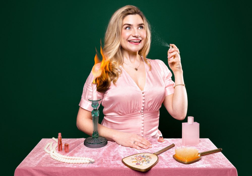 Harriet Kemsley, a white woman with long blonde hair wearing a pink button up blouse, sits behind a table covered with a pink tablecloth. In front of her on the table is a long pearl necklace, a vintage style hairbrush and handheld mirror set, a pink bottle of perfume, and a green candlestick holder holding a lit pink candle, which has singed the ends of Harriet's hair.