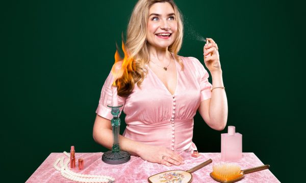 Harriet Kemsley, a white woman with long blonde hair wearing a pink button up blouse, sits behind a table covered with a pink tablecloth. In front of her on the table is a long pearl necklace, a vintage style hairbrush and handheld mirror set, a pink bottle of perfume, and a green candlestick holder holding a lit pink candle, which has singed the ends of Harriet's hair.