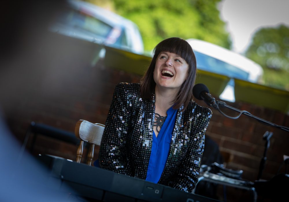 A portrait of singer songwriter Bridie Jackson. Bridie is a white woman, with a shoulder length brown bob and a blunt fringe. She is sitting at a keyboard singing, she is wearing a bright blue blouse and a black, sequinned blazer.