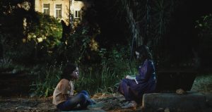 Two young girls are sat outside in the night. One is cross legged on the floor wearing a light t-shirt and jeans with brown mid-length hair, she is facing another girl who is perched on a stone wearing a purple dress with mid-length brown hair. 