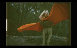 A picture of Anita Pallenberg wearing gold trousers, a dark coloured shirt with a red cape. 
