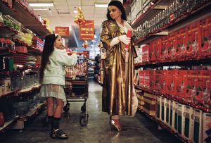 The image shows a shop aisle at Christmas time. It shows a young girl , white, with long dark hair pushing a trolley. She is wearing a white jacket with a white checked skirt. There is a woman to her right white, long dark hair wearing a metallic gold cloak.