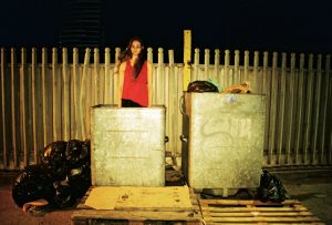 Two industrial-size silver bins in front of a metal fence. A white female is standing in the bin to the left. She has long dark hair and wears a red sleeveless vest. It is night time.