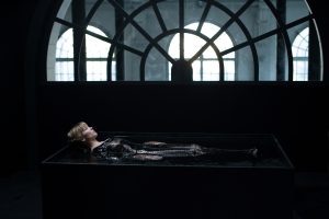 A white female with blonde hair with a fringe in laid horizontal in a pool of dark water. She is enveloped in a black / plastic that covers her full body from her chest. There is a large domed window in the background with white window frames.