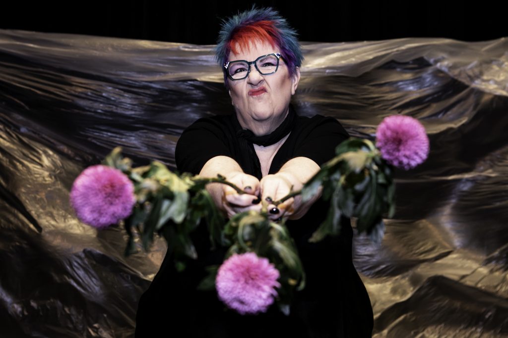 Vici Wreford-Sinnott is an older white woman with pink, blue and purple spiky hair. She is holding three carnation flowers out towards the camera and has a defiant look on her face.