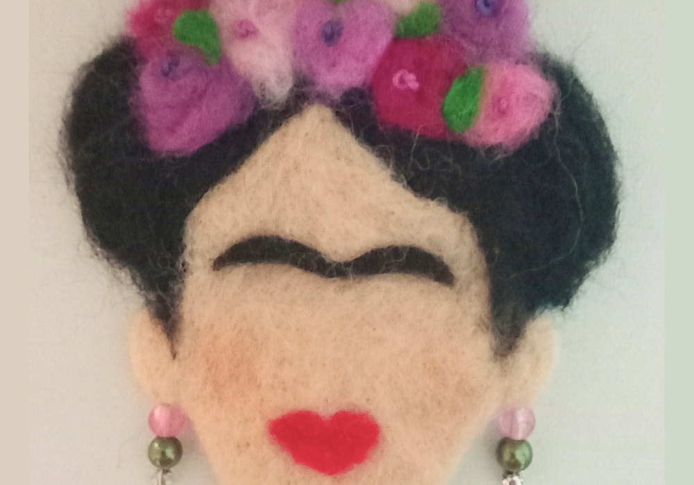A needle felt 2D picture, inspired by Frida Kahlo. It shows a face, missing features like eyes and a nose but with bright red lips and a thick, black brow. The face has black hair, decorated with pink and purple flowers. It is wearing earrings made from pink and green beads.