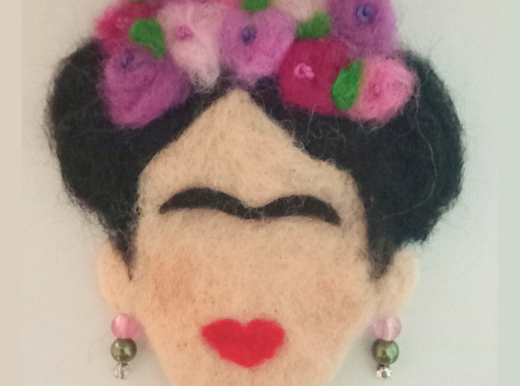 A needle felt 2D picture, inspired by Frida Kahlo. It shows a face, missing features like eyes and a nose but with bright red lips and a thick, black brow. The face has black hair, decorated with pink and purple flowers. It is wearing earrings made from pink and green beads.