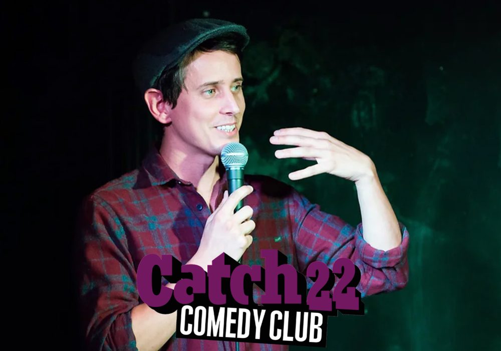 Chris Stokes stands talking into a mic with Catch 22 Comedy Club logo featured.