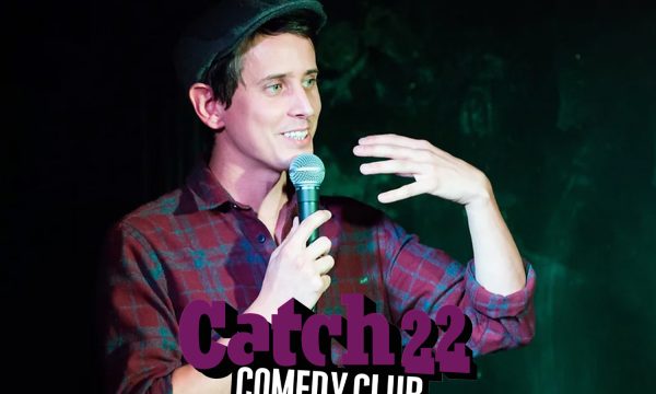 Chris Stokes stands talking into a mic with Catch 22 Comedy Club logo featured.