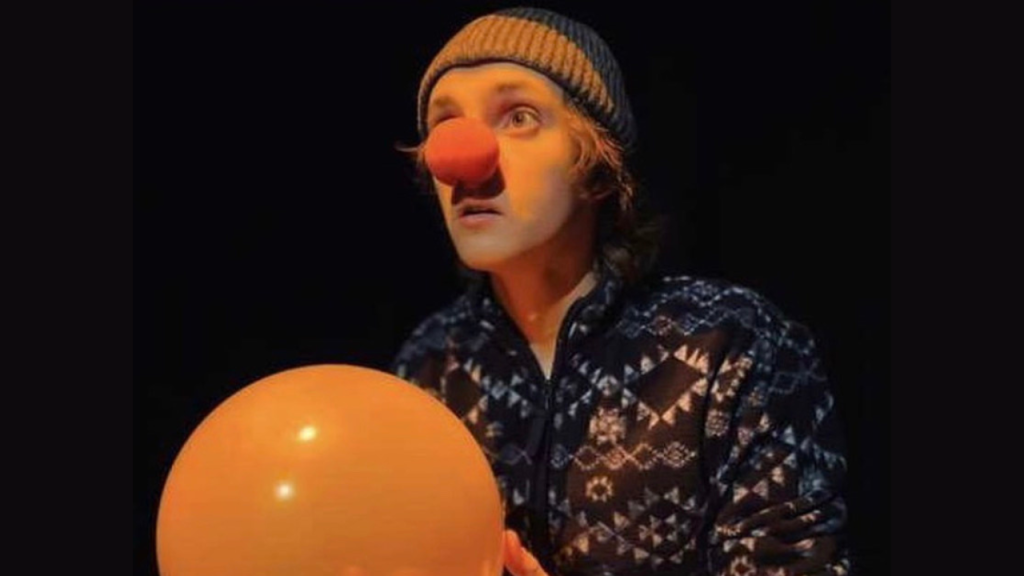 Joseph in character as a clown, wearing a red nose. He is also wearing a wollen beanie hat and has a balloon in his hands.