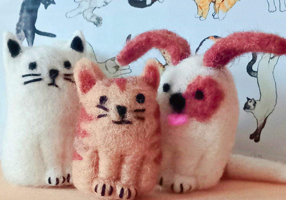 Two needle felt cats and one needle felt dog. The cat on the left is white, with black features, the middle is orange, with dark stripes and black features. The dog on the right is white, with brown floppy ears, a brown patch over one eye, and a pink tongue.