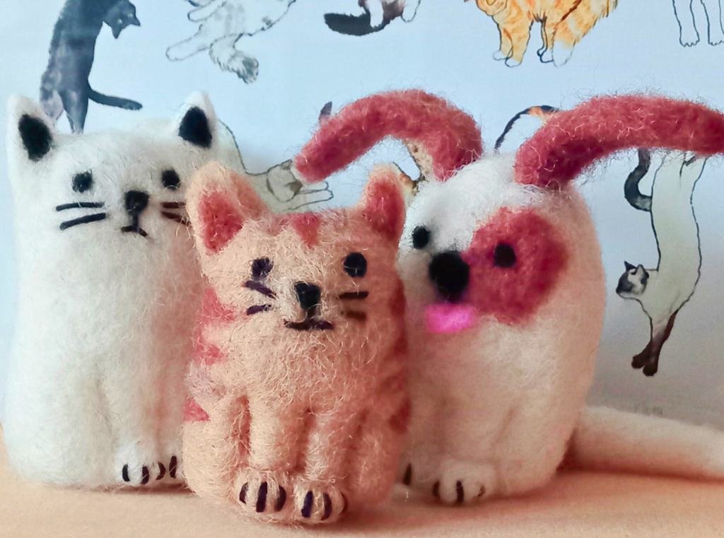 Two needle felt cats and one needle felt dog. The cat on the left is white, with black features, the middle is orange, with dark stripes and black features. The dog on the right is white, with brown floppy ears, a brown patch over one eye, and a pink tongue.