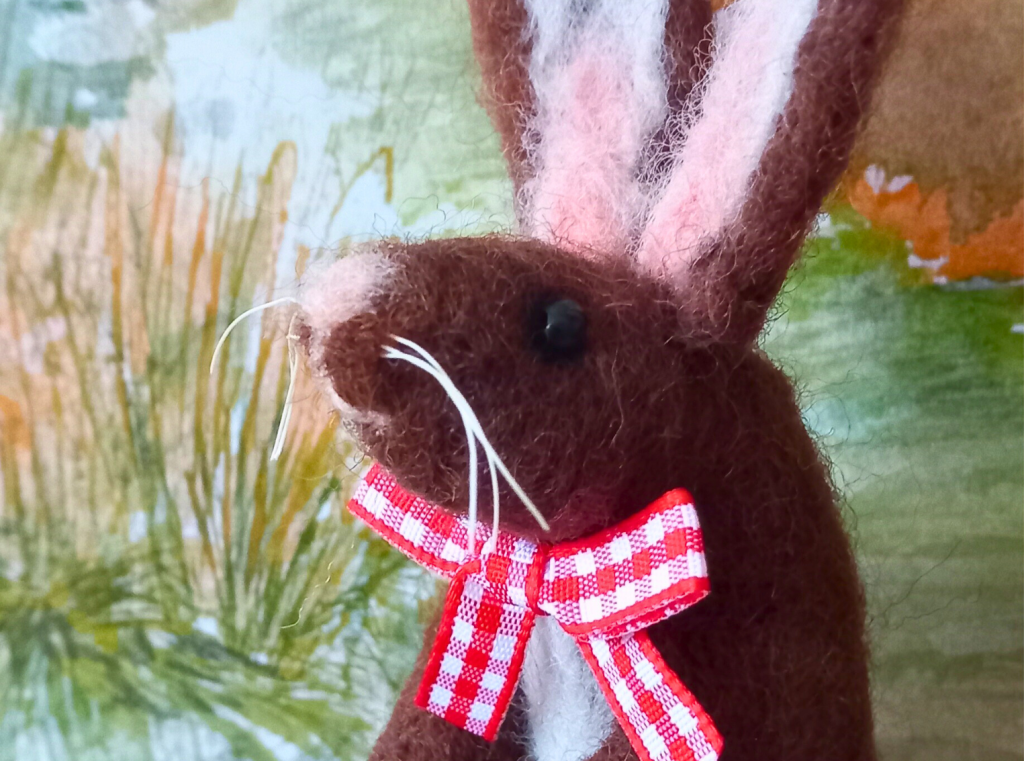 A close up of a needle felted brown hare, it has a pale pink nose and inner ears, and is wearing a red and white gingham bow around it's neck.
