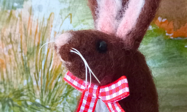 A close up of a needle felted brown hare, it has a pale pink nose and inner ears, and is wearing a red and white gingham bow around it's neck.