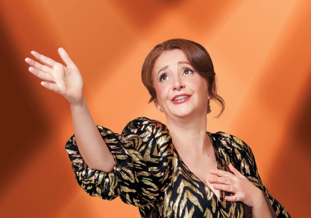 Q white female with mousey-brown hair wearing a black and gold dress. She is holding her chest with one hand and her other hand is raised. Orange background.